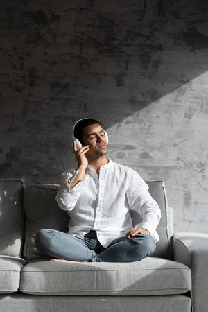 Hispanic young man listening to music on headphones indoors, sitting on sofa in direct sunlight.