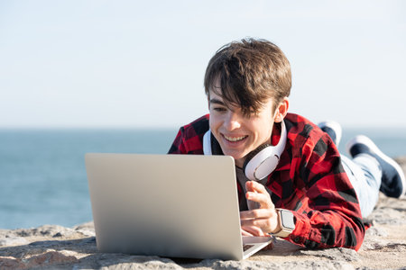 Cute teenager boy talking in front of laptop outdoors. People having video call or live streaming at the coast.の写真素材