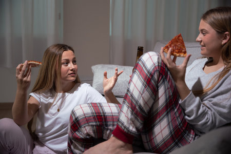 Friends having dinner on sofa. Sisters wearing pajamas eating pizza and talking at night.の写真素材