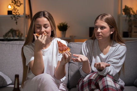 Pretty woman devouring food while friend warning her at night. Sisters wearing pajamas eating pizzaの写真素材