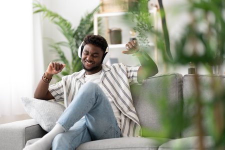 Black young man listening to music and dancing on sofa.の写真素材