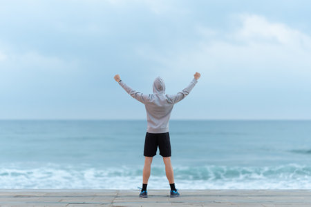 Rear view of sportsman looking at the sea and outstretching arms. Man wearing hoodie celebrating...