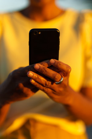 Vertical close-up of the hands of an african woman using smartphone during sunset outdoorsの写真素材