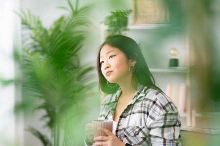 Focus among plants of a dreaming asian woman looking at the window with coffee cup at homeの写真素材
