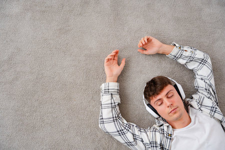 View from above of man listening to music lying on the carpet. Person wearing headphones with closed eyes. Teenager relaxing with copy space.の写真素材