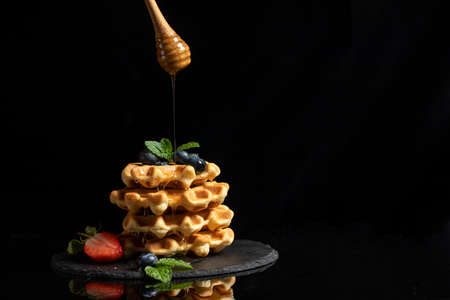 Stack of Belgian waffles with strawberries and mint leaves with pouring honey on black background...
