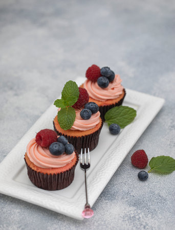 Three mini cupcakes with berry cream blueberry and raspberry mint topping on white plate with fork on a gray background. High quality photoの写真素材