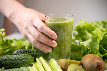 A glass with a green smoothie made from vegetables and fruits. A womans hand holds a glass and wants to drink. Healthy food and the right diet.の写真素材