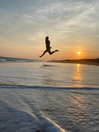 Person captured mid jump above wave on sandy shore of Indian Ocean with dramatic sunset sky, symbolizing freedom, energy, travel, adventureの写真素材