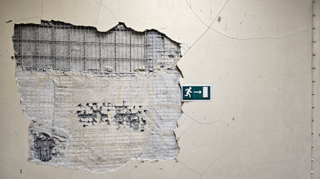 Cracked wall with exposed reinforcement mesh and a green emergency exit sign indicating directionの写真素材