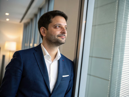 A solitary man stands gazing out a window, conveying a mood of introspection and quiet contemplation. The natural light and blurred background create a serene and thoughtful atmosphere.の素材