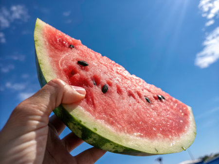 A vibrant red watermelon slice, held delicately by a hand, is showcased against a stunning blue sky filled with fluffy white clouds. The image captures the freshness and deliciousness of summer fruit.の素材