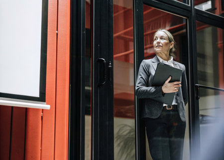 A smartly dressed individual stands in a modern office, poised and focused, holding a folderの素材