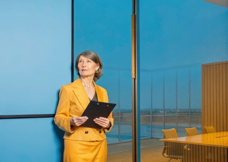 A stylish individual, clad in a bright yellow blazer and dark skirt, concentrates on work within a bright, modern conference room. Natural light and a professional design create a sense of clarity and focus.の素材