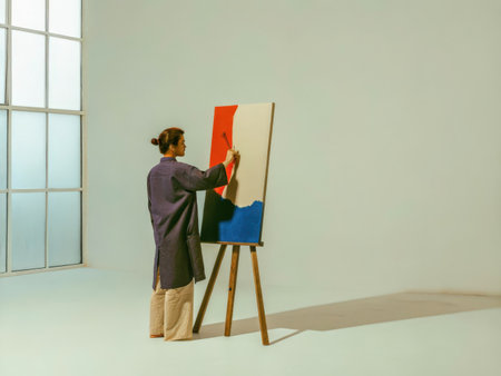 A focused artist stands before a large canvas, diligently applying vibrant colors to an abstract design. Soft window light illuminates the scene, capturing a moment of creative concentration.の素材