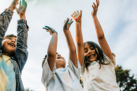 Three children, covered in paint, reach upwards towards a bright blue sky, conveying a sense of wonder and playful excitement. The blurred background enhances the feeling of movement and youthful energy.の素材