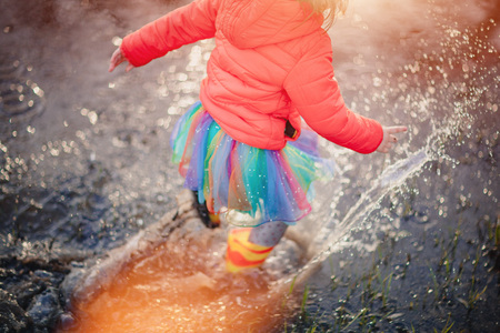 Crop view from above of girl in colorful skirt and gumboots jumping in big puddle splashing water...