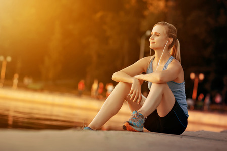 Young beautiful girl sitting on the edge of the city lake dam and listens to music player.の写真素材