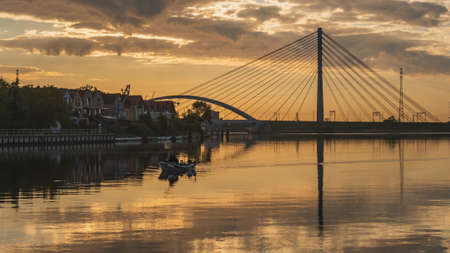 Suspension bridge over the Martwa WisÅa in GdaÅsk at sunset and a fisherman in a small boat. Cool atmosphere gilded with beautiful light.の写真素材