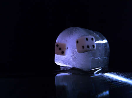 two playing dice are frozen to a cold piece of ice with bubbles, which lies on a vinyl record on a black background and is highlighted with a blue backlight in a macro photoの写真素材