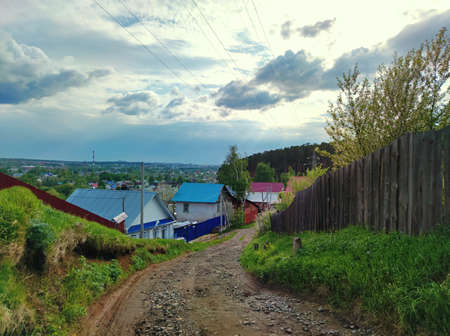 a rural road strewn with stones leads near the fence to the village against the sky with clouds before the rainの写真素材