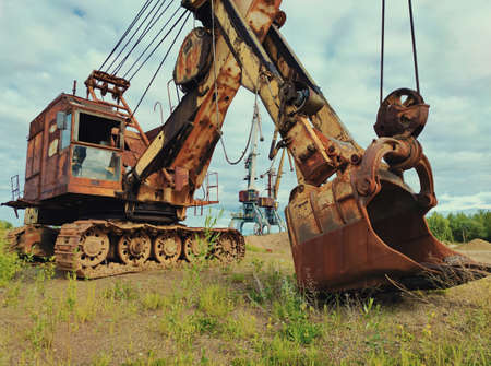 old huge excavator with peeling paint and tall cranes on a sand pit against a beautiful blue cloudy...