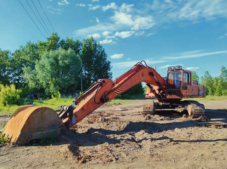 old excavator with a huge bucket on a sandy platform against a background of green trees and a...