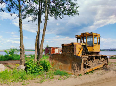 old yellow bulldozer stands near green trees near the riverbank against a beautiful blue sky with cloudsの写真素材