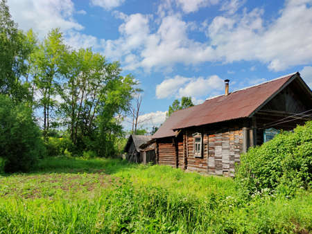 green courtyard near an old wooden house and trees against a blue sky with clouds on a sunny dayの写真素材