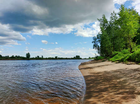 gloomy clouds in the blue sky over the sandy bank of the river with green trees