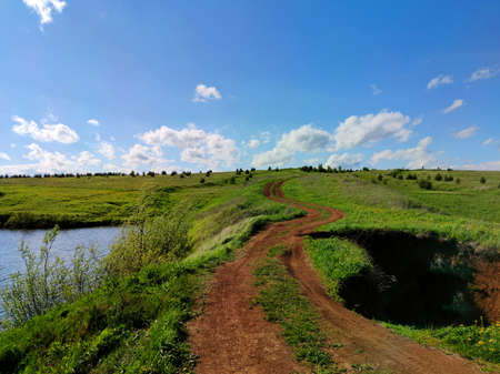 rural winding road uphill through a field between a lake and a ravine against a blue sky with clouds...