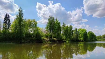 green trees by the lake are reflected in the water against a blue sky with clouds on a sunny day