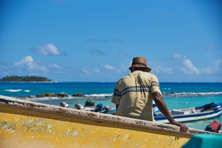 fisherman sitting in his wooden boat that is on the beach with a blue sea with waves in the background. San Andres Island, Colombia. December 11, 2020.のeditorial素材