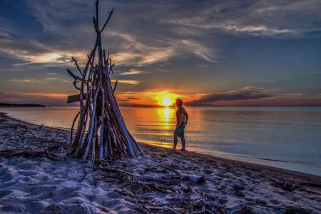 Sunset Reflections  Teenaged male on the beach with a handmade shelter out of driftwood watching the...