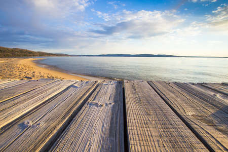 Summer Vacation At The Beach. Wooden dock overlooks a sunny sandy beach with a blue water horizon...
