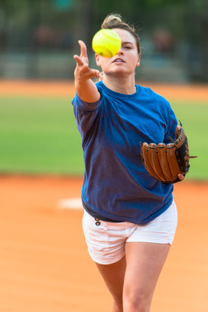 Young woman softball player pitching fluorescent yellow softball on field.