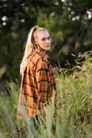 Young woman standing in tall grass field wearing a plaid shirt and looking over her shoulder at...