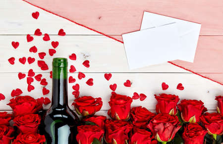 Buds of red roses and bottle of wine on a white wooden background with confetti and messageの写真素材
