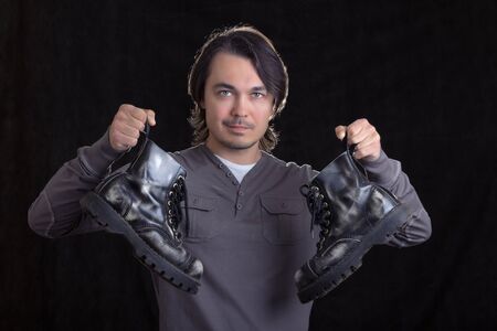 Young man holding old black boots in his hands, on black background