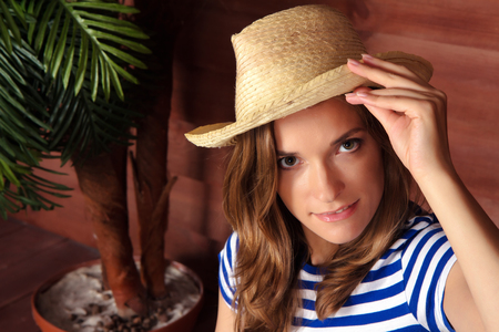 Beautiful woman in a striped shirt holding her straw hat and looking at a camera. Wooden veranda of a summer country house sa a backgroundの写真素材