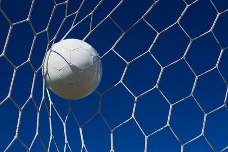Close-up of a soccer ball (football) going into the back of the net with a blue sky background.の写真素材