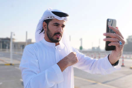 Middle Eastern Emirati Arab model fixing his traditional menswear garment Ghutra using his phoneの写真素材