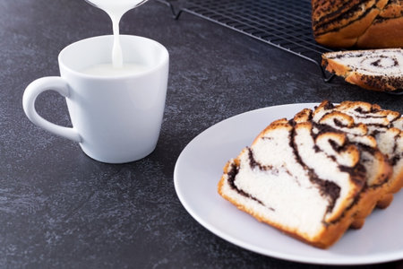 Milk is poured into a mug, next to a bun with poppy seed filling on a white plate, dark background