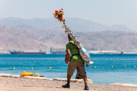 Eilat, Israel - May 15, 2012: Man with red hat caring authentic tea vessel container on his back,...