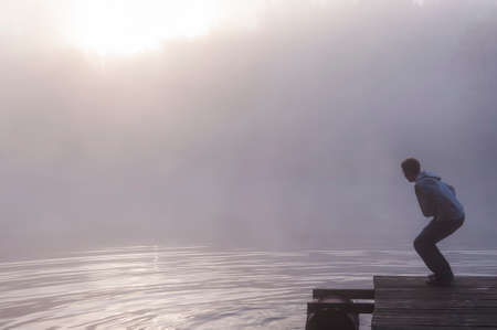 Young man making water ripples at foggy sunrise by jumping on lake pier