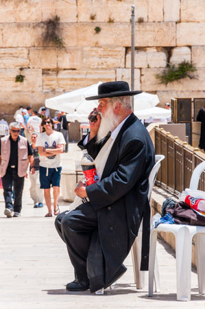 Jerusalem, Israel - May 25, 2012: Jewish orthodox in black suit and hat sitting and drinking near the entrance alley to the Western Wall. People, locals and tourists walking around the plazaのeditorial素材