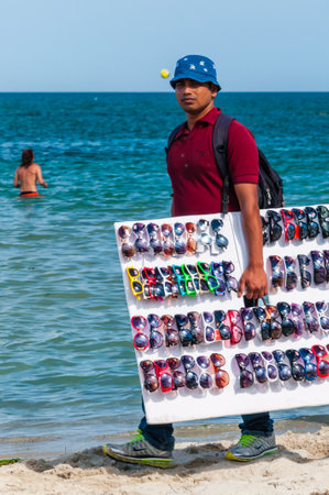 Paralia, Greece - June 14, 2013: Man holding showing offering selling various sunglasses while walking on the beach between resting tourists. Seasonal beach resorts businessesのeditorial素材