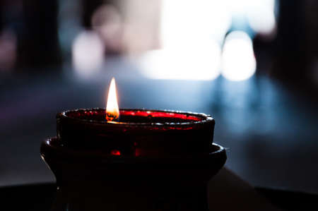 Red glass burning candle close-up in dark interior with light on the background