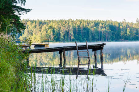 Old wooden pier with ladder on foggy lake with amazing forest on background at sunriseの写真素材