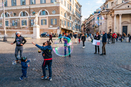 Rome, Italy - November 18, 2018: Kids catching and blowing soap bubbles flying on Piazza del Popolo, People Square in Rome full of people, tourists and locals with Roman architecture on backgroundのeditorial素材